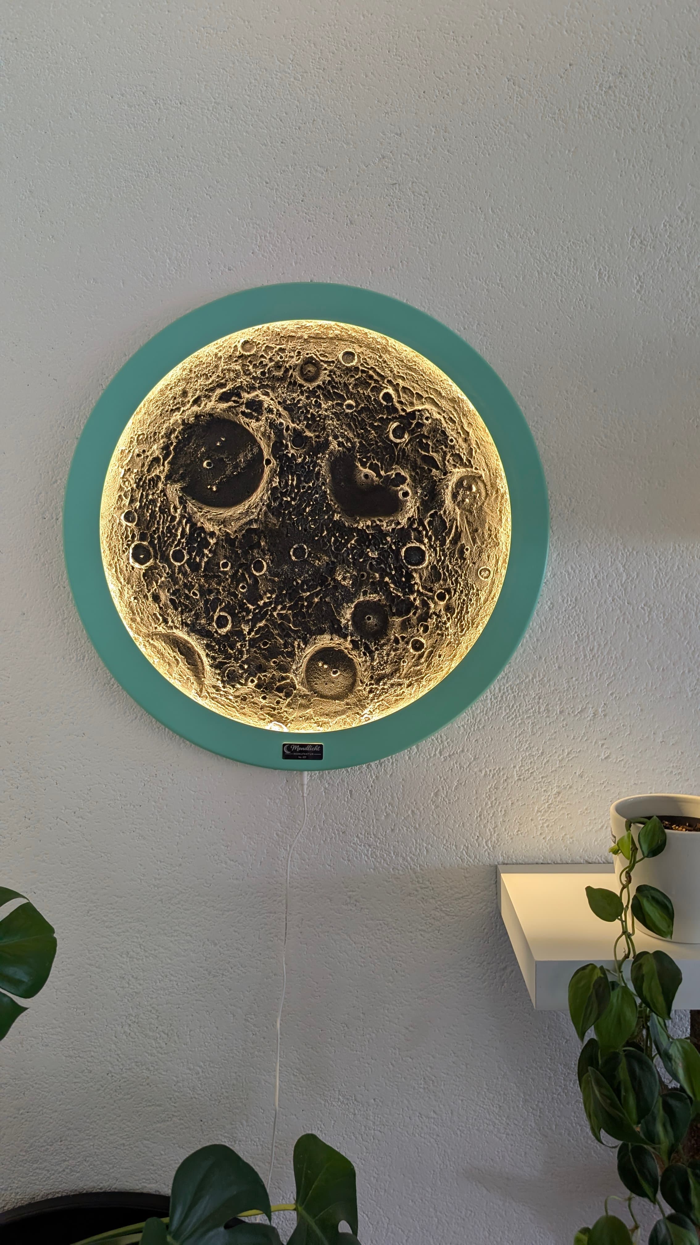 Eine leuchtende, kreisförmige Mondlampe mit detaillierten Kratern und türkisfarbenem Rahmen an einer weißen Wand.